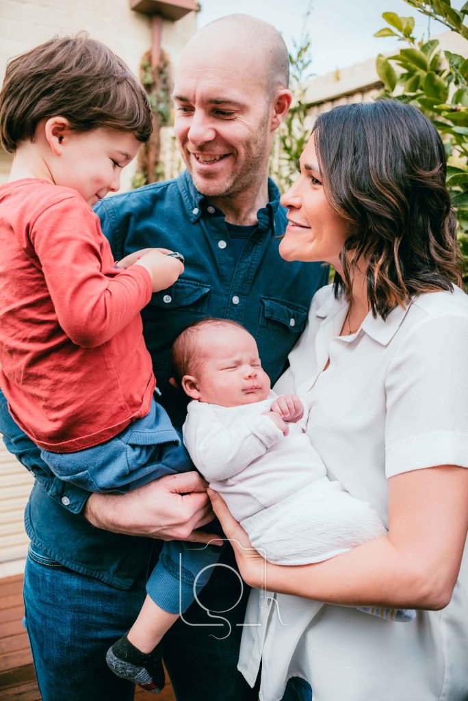 A lifestyle photography session with a Melbourne family in their backyard. A father holding and looking at his son who is holding Lego. A mother holding her newborn baby girl looking in the direction of her son. Family Photography by Liyat Haile.