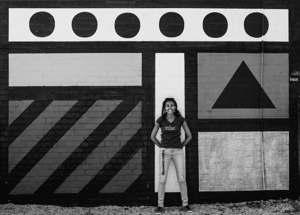 Liyat Haile Photographer standing in front of a wall with shapes. Liyat is looking straight into camera and smiling with her hands behind her.